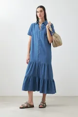 Maxi vestido camisero de jean azul con cuello clásico, manga corta, cierre frontal con botones y falda amplia con volantes.