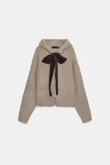 Chaqueta de punto con capucha, manga larga y cierre frontal con cremallera. Presenta un diseño en color beige con un lazo decorativo a contraste en color marrón oscuro en la zona del cuello.