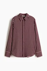 Camisa de manga larga con cuello clásico, cierre frontal abotonado y puños con botones. Presenta un diseño de rayas verticales en tonos bordó y crema, con bajo redondeado.