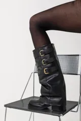 Botas de caña alta confeccionadas en cuero negro, con diseño holgado y tres correas horizontales con hebillas metálicas doradas en la parte superior. Presentan punta redondeada y taco bajo cuadrado.