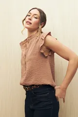Camisa color beige con textura suave, cuello mao con volado y mangas cortas con pequeño volado. Presenta botonadura frontal.