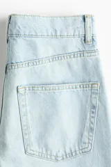 Jean de tiro alto confeccionado en denim de algodón rígido, con diseño curvo y voluminoso en las piernas, tipo barrel. Presenta cinco bolsillos, cierre frontal con botón y detalles de tiras para anudar en los tobillos.