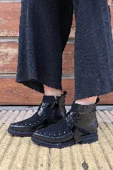 Bota corta de cuero y gamuza color negro, con taco bajo y suela plana. Presenta una correa lateral de cuero charolado con hebilla metálica, tachas decorativas en la puntera y paneles elásticos en los laterales de la caña.