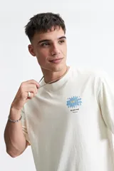 Remera de corte oversize color crudo, con cuello redondo y mangas cortas. Presenta un estampado frontal con diseño de flor azul y texto.