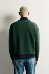 Chaqueta regular fit color verde oscuro con detalles en negro en el cuello subido, los puños, el dobladillo inferior y los ribetes de los bolsillos. Cuenta con cierre frontal de cremallera metálica y bolsillos con cierre en la cadera.