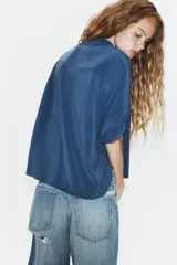Camisa de corte fluido con cuello solapa y manga corta con dobladillo. Presenta un cierre frontal con botones ocultos bajo una tapeta y un diseño con bajo ligeramente más largo en la parte posterior.