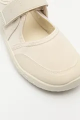 Zapatilla tipo guillermina de estilo barefoot, color beige con detalles en rosa pálido en el talón y tira de ajuste con velcro. Posee suela plana y flexible diseñada para un movimiento natural del pie.