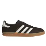 Championes Adidas Gazelle Indoor, modelo de corte clásico y retro fabricado en gamuza de color marrón oscuro, con las tres franjas características de la marca y la lengüeta del talón en color blanco. Incluye la inscripción "GAZELLE" en letras doradas en el lateral y suela de caucho translúcido tipo caramelo.