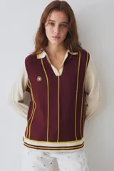 Remera tejida de punto estilo polo con mangas largas en color crema y cuerpo en color bordó con rayas verticales amarillas. Presenta cuello tipo polo, aberturas laterales y un pequeño parche circular con diseño de sol en el pecho.