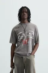 Camiseta gris de punto con efecto lavado y roturas, corte "boxy fit", cuello redondo y manga corta. Presenta un estampado frontal en blanco y rojo con diseño abstracto.