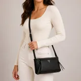 Cartera Guess modelo Devon de color negro, confeccionada en material sintético con textura granulada. Presenta un diseño rectangular, herrajes metálicos dorados, logo de la marca en la parte frontal y un charm colgante decorativo. Cuenta con cierre superior y una correa ajustable para llevar al hombro o cruzada.