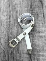 Cinto blanco de cuero sintético con hebilla metálica labrada y ojalillos metálicos.