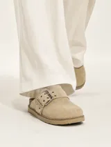 Zueco de gamuza color beige, con diseño de punta cerrada y talón descubierto. Presenta una tira ajustable sobre el empeine con hebilla metálica y ojales decorativos. Cuenta con una suela de corcho y base de goma antideslizante.
