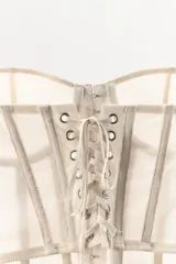 Corsé strapless color beige claro, con copas con aros y estructura de varillas verticales. El tejido es semitransparente con pequeños lunares visibles. Presenta un cierre frontal con ganchos.
