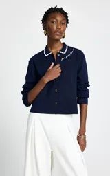 Cardigan de punto azul marino con cuello polo, cierre frontal de botones y detalle de bordado en el pecho. El cuello presenta un borde festoneado en color blanco.