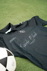 Camiseta de fútbol estilo boxy de manga corta y cuello polo, en color negro con sutiles rayas verticales. Presenta inscripciones estampadas y escudo bordado en el pecho.