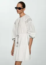 Vestido corto de algodón blanco con detalles bordados en negro en hombros y mangas. Presenta cuello redondo con abertura frontal, mangas largas abullonadas con puños elásticos y cintura elástica ajustable con lazo decorativo con borlas.