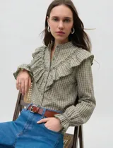 Camisa de manga larga con estampado de cuadros vichy en tonos verde oliva y crudo. Presenta cuello mao, cierre frontal con botones y volados decorativos en la parte delantera. Las mangas terminan en puños elastizados con un ligero acabado de volado.