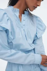 Vestido corto de algodón celeste, con cuello alto con volados y abertura frontal con botones. Presenta mangas largas, elástico en la cintura y falda escalonada con detalles de encaje y volados en los bordes.