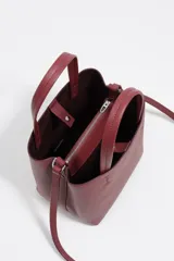 Bolso pequeño de piel color borgoña, con estructura rígida y forma de cubo. Posee doble asa de mano y una correa larga para llevar al hombro o cruzado. El frente lleva el logo metálico de Bimba y Lola.