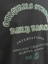 Buzo de manga larga y cuello redondo, color negro, con fit holgado. Presenta un estampado frontal en tonos verde menta y blanco con texto que incluye "Originals Studio", "Daily Basics", "International", "connecting CREATIVITY ACROSS DISTANCES" y "est. 90".