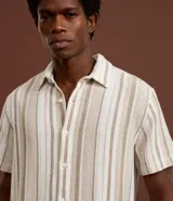 Camisa de hombre de manga corta con cuello tradicional y abotonado frontal. Presenta un tejido texturizado con un patrón de rayas verticales en tonos beige, blanco y marrón claro.