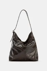 Bolso de hombro tipo shopper, color marrón oscuro con efecto envejecido o desgastado. Presenta un asa de hombro con detalle de tachas metálicas y ajuste mediante hebilla. El cierre principal es magnético e incluye un bolsillo interior con cremallera.