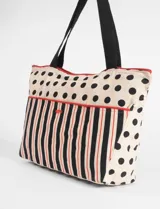Bolso grande de tela con diseño combinado de lunares negros sobre fondo crema y bolsillo frontal con estampado de rayas verticales negras y rojas. Cuenta con ribetes en color rojo y correas negras.