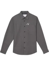 Camisa de manga larga color gris oscuro, con cuello clásico, bolsillo de parche en el pecho izquierdo y botones oscuros. Presenta un pequeño logo estampado en blanco en el bolsillo y una etiqueta con el texto "Calvin Klein Jeans" en el cuello.
