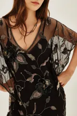 Vestido corto de tul negro con transparencias, bordado con lentejuelas y motivos florales. Presenta mangas cortas tipo kimono, escote en V y un cinturón de tela con flecos y detalles brillantes. Incluye un viso interior.