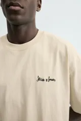 Camiseta relaxed fit color beige, con cuello redondo y manga corta. Presenta un bordado y estampado combinado a contraste en el delantero y la espalda.