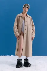 Trench midi de color camel, confeccionado en una mezcla de algodón y nylon con propiedades repelentes al agua. Presenta un diseño clásico con cuello camisero, cierre frontal de botones, bolsillos laterales y tajos en los costados.