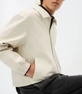 Campera masculina de corte boxy en color beige, con cuello inglés, cierre frontal de cremallera, mangas largas con puños elásticos y ruedo con elástico. Presenta bolsillos laterales y detalles de recortes en el diseño.