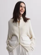 Cárdigan tejido color crema con cierre frontal de cremallera dorada, cuello alto y mangas largas raglán. Presenta dos bolsillos frontales de cuero sintético con solapa y botón a presión.