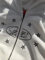 Campera canguro blanca con cierre frontal, capucha y bolsillos tipo kankaro. Presenta un estampado frontal con un óvalo con texto cursiva rodeado de estrellas negras.
