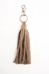 Charm para bolso o llavero compuesto por flecos de cuero sintético en color beige, decorados con pequeñas tachas metálicas plateadas. Incluye una argolla y un cierre de mosquetón metálico.