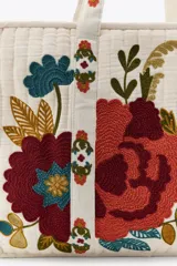 Bolso tote acolchado de color blanco crudo, con asas de hombro y bordados florales grandes y coloridos en tonos rojo, marrón, azul y verde. Presenta un forro interior y cierre de cremallera metálica.