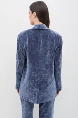 Blazer de terciopelo azul con solapas anchas y cierre frontal de un botón. Presenta un diseño entallado de manga larga con textura suave y acabado ligeramente brillante.