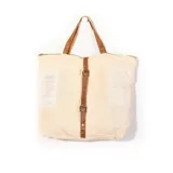 Cartera tipo tote de yute color beige, con asas de hombro, detalles de tiras de cuero marrón con hebillas metálicas y cierre superior.