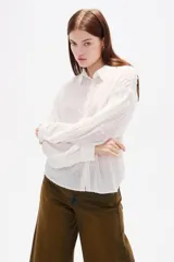 Camisa de color blanco con textura de pequeños puntos en relieve, cuello clásico, cierre frontal con botones y mangas largas con detalle de punto smock y puños elásticos.