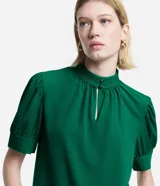 Blusa verde esmeralda de crepé con cuello alto con botones dobles y pequeña abertura abajo y mangas cortas abullonadas con botones.