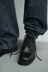 Bota de cuero negro con cordones tipo derby, parte superior lisa y plantilla interior de piel. Presenta horma cuadrada y suela con ligero tacón.