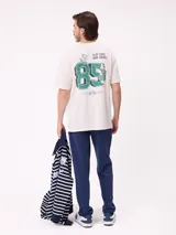 Remera de corte holgado color crema con estampado gráfico en la espalda que incluye el número 85, personajes animados y la frase Play Hard, Surf Harder.