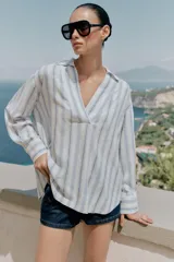 Blusa de manga larga confeccionada en mezcla de viscosa, lino y liocel, con diseño de rayas verticales en tonos celestes y marrones sobre fondo blanco. Presenta cuello solapa con escote en V cruzado.