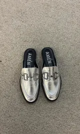 Suecos tipo mocasín de cuero metalizado plateado, con detalle de estribo metálico en el empeine y suela plana negra.