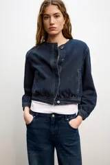 Chaqueta estilo bomber confeccionada en tejido denim de color azul oscuro. Presenta cuello redondo, manga larga, bolsillos delanteros de vivo y cierre frontal con botones ocultos.