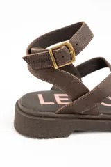Sandalias planas de cuero marrón con tiras cruzadas, sujetadedos y tira con hebilla ajustable al tobillo.