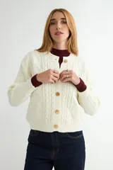 Cardigan de punto trenzado color crema con cuello redondo y cierre frontal de botones grandes en tono dorado. Presenta mangas largas con puños acanalados y un diseño de tejido con textura.