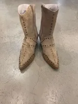 Bota de caña media color beige de gamuza, con punta afilada y taco bajo. Presenta detalles de tachas metálicas doradas a lo largo de la caña y el contorno de la puntera. Posee cierre lateral.