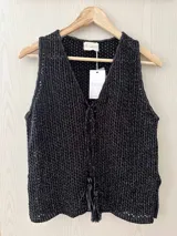 Chaleco tejido en punto calado con hilo de algodón y lurex, de color negro con destellos plateados. Presenta escote profundo en V, cierre frontal con cordones entrelazados terminados en borlas y un corte recto y holgado.